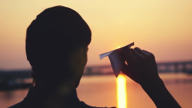 Young guy launch paper plane against the sea during sunset with sun flare and reflections in the water, as in childhood in slowmotion. 1920x1080