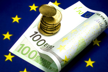One hundred euro bill lying in the center of the star circle on the European community flag. Several euro cents coins on the bill. Macro image.
