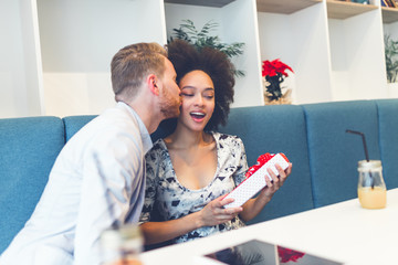 Happy middle age interracial couple sitting in cafe bar. Man giving Valentine's day gift to his girlfriend or wife. 