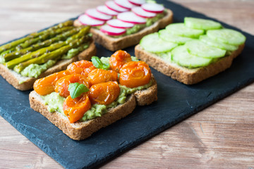 Selection of vegetarian toasts sandwiches made with baked tomoatoes, cucumbers,  asparagus, radish, blackberries on the black platter, selective focus