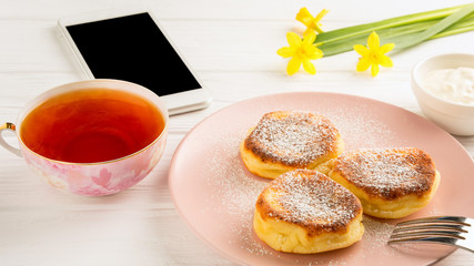  Cheese pancake with powdered sugar black tea flowers and smartphone on a white wooden table.