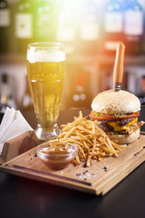 Tasty grilled cheeseburger with a knife stabbed trough on a wooden plate. Napkins and beer on the side. Lens flare in the background.