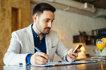 Young businessman taking note and looking at phone in cafe