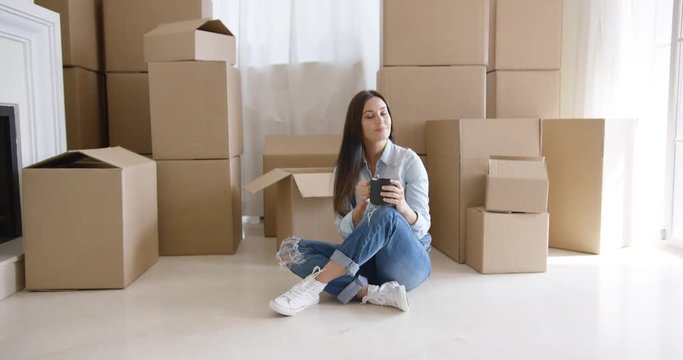 Young woman relaxing on the floor of her house with a mug of coffee surrounded by brown cardboard boxes for moving