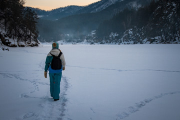 The girl with a backpack walking on the frozen river against the backdrop of rugged mountains