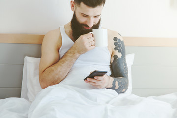 Bearded man lying in bed with morning coffee and phone using app