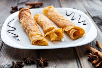 Sweet fresh homemade crepes with chocolate topping on dark grey rustic wooden table. Menu for breakfast, desserts and lunch. Cinnamon and kitchen herb for decoration.
