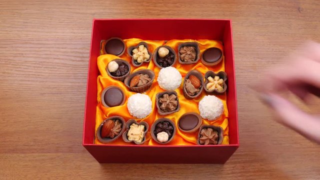 A box of chocolates on the table. One opens the box with candies. Many hands taking candy from a box