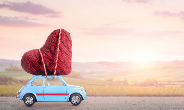 Blue retro toy car delivering heart for Valentine's day against blurred sunset rural landscape