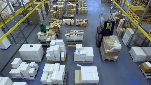 Worker in loader transporting goods cardboard boxes in warehouse storage HD video. Forklift rides between shelves. Man working on lift truck