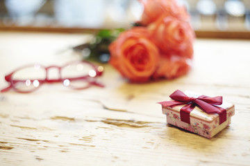 Small present box with bow on wooden table. Focus on bow. Red roses flowers behind on wooden table. St. Valentine's day concept. Blank space for text