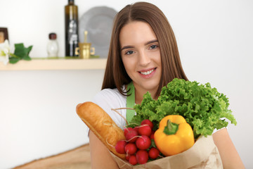 Young caucasian woman in a green apron is holding paper bag full of vegetables and fruits while smiling in kitchen