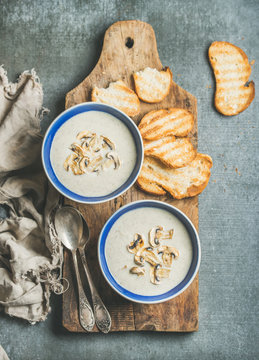 Creamy mushroom soup in bowls with toasted bread slices on rustic serving board over grey concrete background, top view