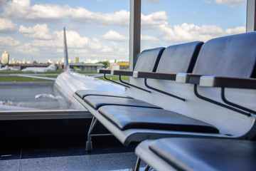 Row of empty black leather seats in waiting lounge at the airport on the background of viewing glass, blurry airplanes, buildings, white clouds and blue sky at sunny morning