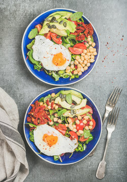 Healthy breakfast with fried egg, chickpea sprouts, seeds, fresh vegetables and greens in blue bowls over grey concrete background, top view. Clean eating, healthy lifestyle, vegetarian food concept