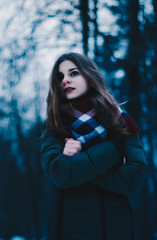 Portrait of a beautiful brown-haired young girl, with pale face, red lips, in a jacket and a scarf tied around the neck, on the background of dark trees. Portrait of a beautiful girl.