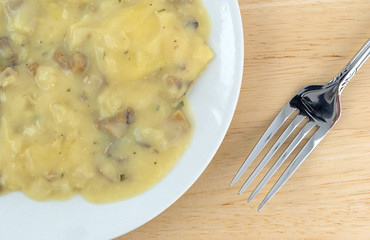 Top close view of ravioli in a cheese and mushroom sauce meal on a white plate with a fork to the side atop a wood table.