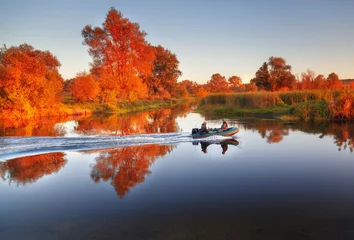 Fototapeten Fishermen sailing fast at rubber powerboat by river making trace at tranquil water surface against beautiful morning scenery with golden color foliage trees at bank. Fishing background. © Feel good studio