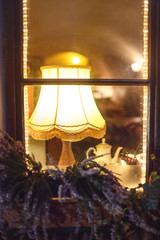 Window frame from outside, decorated with flowers, and a lamp, warm light and a tea pot inside the house.