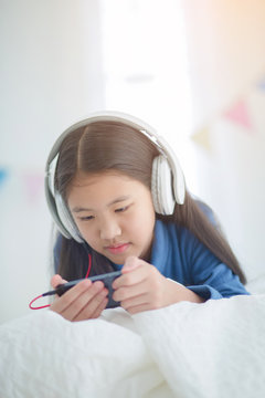 Pretty Asian girl using headphone for listen music by smartphone on the bed in her decorated bedroom