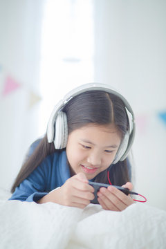 Pretty Asian girl using headphone for listen music by smartphone on the bed in her decorated bedroom