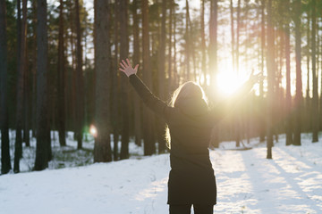 teen girl raised hands from behind in winter pine forest in sunset, freedom or victory concept