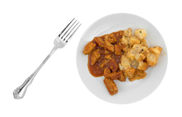 Top view of  a plate with chicken chunks in barbecue sauce plus potatoes and a fork to the side isolated on a white background.