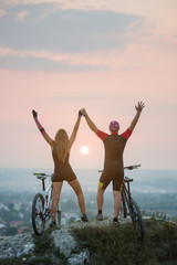 Magnificent sunset, bright sun between cyclists on the cliff top. Silhouette of guy and girl on mountain bikes keep the hands lifted upwards. Pink Kinesio tape glued on the girl's hand