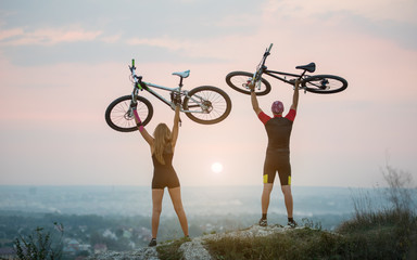 Bikers holding bikes high up in the sky on the top of a hill against magnificent sunset with blurred background. Pink Kinesio tape glued on the girl's hand.