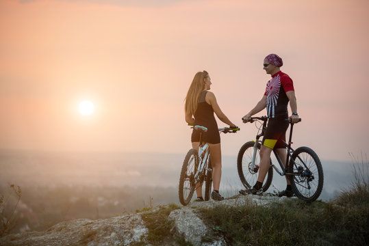 Rear view cyclist couple with mountain bikes standing on a rock, enjoying the evening scenery, looking to each other. Blurred background