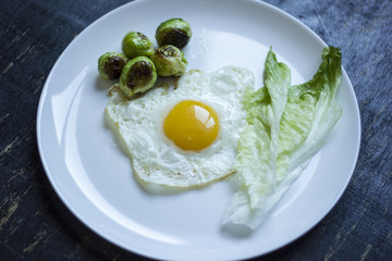 fried egg with cucumbers, olives, and brussels sprouts on a plate with fork and knife on a dark wooden background