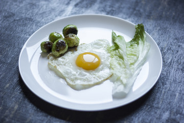 fried egg with cucumbers, olives, and brussels sprouts on a plate with fork and knife on a dark wooden background
