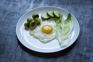 fried egg with cucumbers, olives, and brussels sprouts on a plate with fork and knife on a dark wooden background