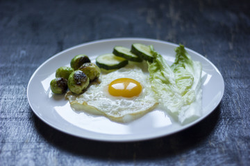 fried egg with cucumbers, olives, and brussels sprouts on a plate with fork and knife on a dark wooden background