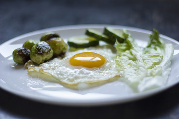 fried egg with cucumbers, olives, and brussels sprouts on a plate with fork and knife on a dark wooden background