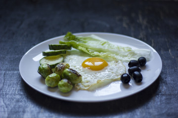 fried egg with cucumbers, olives, and brussels sprouts on a plate with fork and knife on a dark wooden background