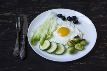 fried egg with cucumbers, olives, and brussels sprouts on a plate with fork and knife on a dark wooden background