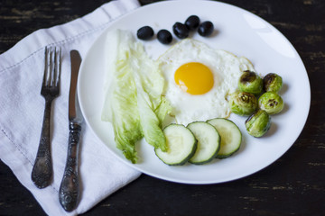 fried egg with cucumbers, olives, and brussels sprouts on a plate with fork and knife on a dark wooden background