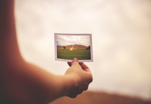 a woman holding an instant photo of a hill in front of her - des