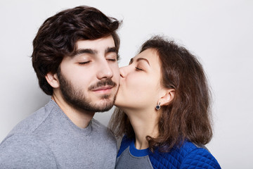 Romantic loving couple. A pretty brunette kissing his bearded boyfriend in his chin. A happy couple closing their eyes from pleasure isolated over white background having romantic evening