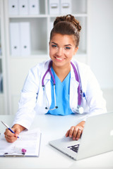 Beautiful young smiling female doctor sitting at the desk and writing.