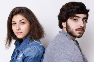 Two young people sitting back to back looking directly into camera. A young couple: brunette girl in jean clothes and stylish bearded man in grey sweater sitting backs relaxing