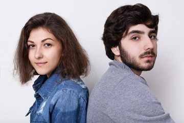 A bristly man and brunette woman sitting back to back relaxing at home looking into the camera with serious expression. Young hipsters sitting close to each other. Love and relationships concept