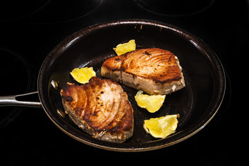 Two tuna steaks and lemon pieces in a frying pan on a black stove