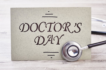 Stethoscope near Doctor's Day card. Greeting paper and wooden background. Stressful and honorable profession.