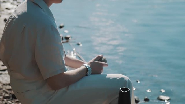 Man in hair net and white shirt sitting at lake shore pretending to throw ston into camera on summer sunny day