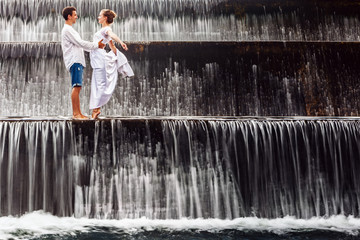 Fototapeta premium Happy family on honeymoon holidays - married loving couple hugging, kissing with fun under falling water in cascade waterfall pool. Active lifestyle, people travel on summer vacation on Bali island.