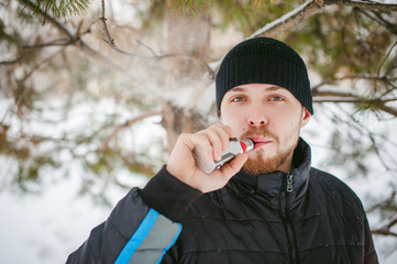 a young man with a beard, smokes an electronic cigarette, outdoors in winter snow