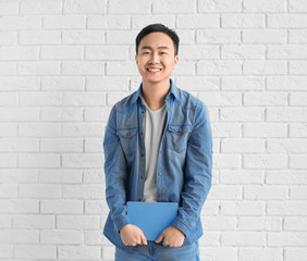 Young Asian man with book standing near brick wall