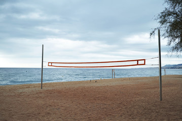Fototapeta na wymiar Landscape view on empty morning beach with volleyball grid stretched between two steel poles, dramatic cloudy sky and blue sea behind